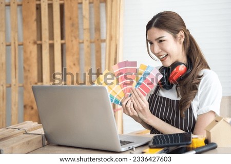 Happy female carpenter holding pantone in hand in her workshop. She looking to laptop with smiling in her workshop.