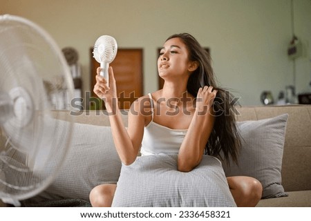 A gorgeous Asian woman, feeling hot and tired, refreshes herself with a handy fan and an electric fan while sitting on a couch in her living room on a summer day. live without air condition Royalty-Free Stock Photo #2336458321