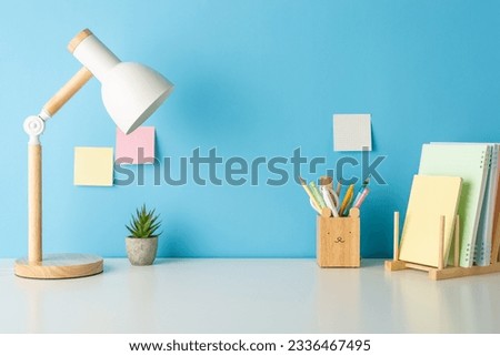 Education-themed composition: Side view photo of desk arrangement displaying pencil holder, pens, books stand. Lamp and plant enhance scene. Blue wall backdrop with sticky notes offers space for text