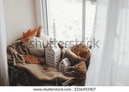 Cozy Autumn. Stylish pumpkin pillows, fall leaves, candle, lights and cute buildings decoration on brown scarf on windowsill. Autumn hygge, fall home decor. Happy Thanksgiving