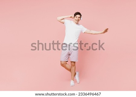 Full body smiling happy fun young caucasian man wearing white t-shirt casual clothes listening to music in headphones isolated on plain pastel light pink background studio portrait. Lifestyle concept
