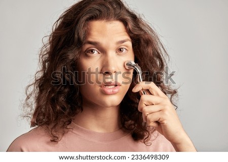 Young handsome long haired guy using de-puffing face roller to keep his skin soft and smooth. Caucasian man with brown hair maintaining his natural beauty with skincare routine. Men's self-care.