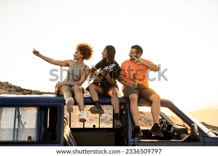 A group of 3 young multi-ethnic people are having fun on top of a camper van at sunset. Friends are taking a picture with the mobile phone while drinking beer and playing a flamenco guitar.