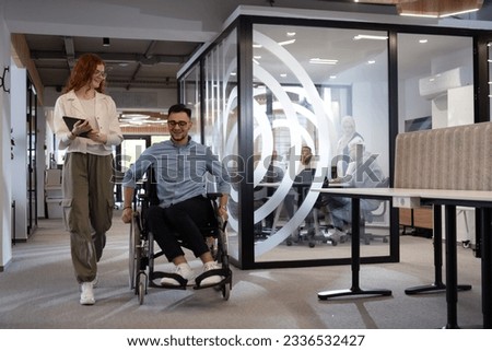 Young business colleagues, collaborative business colleagues, including a person in a wheelchair, walk past a modern glass office corridor, illustrating diversity, teamwork and empowerment in the Royalty-Free Stock Photo #2336532427