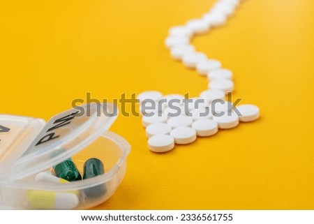 Arrow shaped white medical pills point to the plastic daily organizer (for a week of medication) and flow towards it. Close-up on a yellow background.