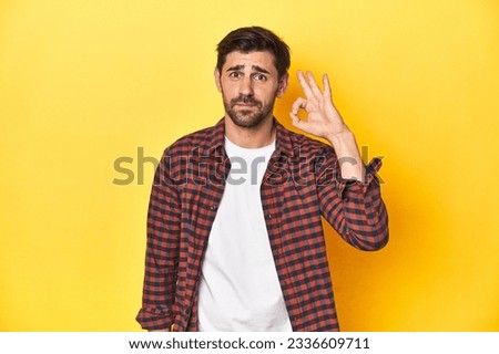Caucasian man in red checkered shirt, yellow backdrop cheerful and confident showing ok gesture.
