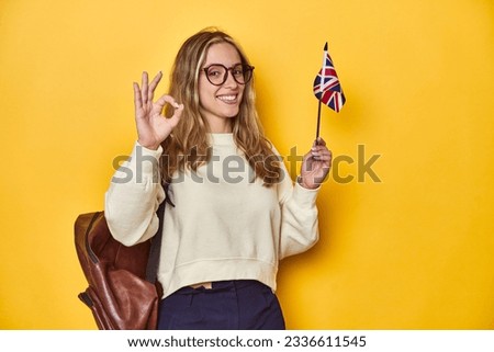 UK flag-waving student, Erasmus concept on yellow backdrop, cheerful and confident showing ok gesture. Royalty-Free Stock Photo #2336611545