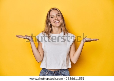 Young blonde Caucasian woman in a white t-shirt on a yellow studio background, makes scale with arms, feels happy and confident.