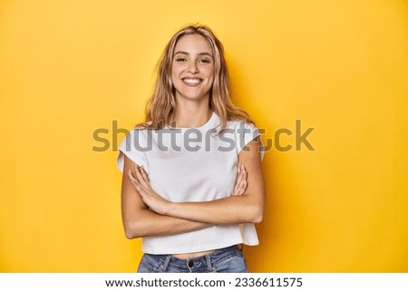 Young blonde Caucasian woman in a white t-shirt on a yellow studio background, who feels confident, crossing arms with determination.