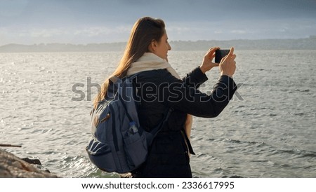 Young woman with backpack on the rocky coast, taking photos of the cold winter sea on her smartphone. Perfect for tourism and travel videos