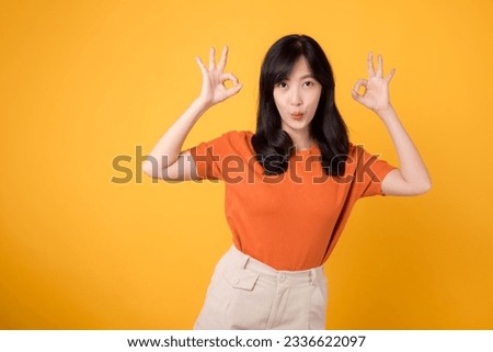 In a vibrant yellow backdrop, a young Asian woman 30s, donning an orange shirt, displays the okay sign gesture. Hands gesture concept.