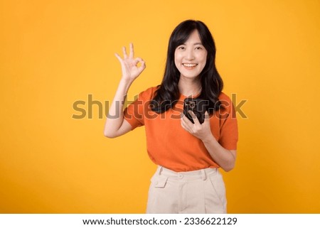 Spread happiness with a young Asian woman 30s, donning an orange shirt, showing okay hand sign while using smartphone on yellow background.
