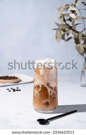 Iced coffee latte in a tall glass with milk on a light background with coffee beans, spoon and morning shadows. Summer refreshment concept. Front view.