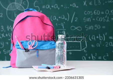 Colorful school backpack with bottle of water and stationery on white table near green chalkboard Royalty-Free Stock Photo #2336631305