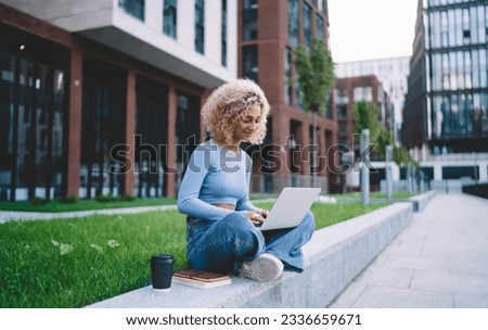 Full body of smiling young woman in casual clothes with curly hair sitting on parapet with crossed legs and studying online using laptop while looking at screen Royalty-Free Stock Photo #2336659671