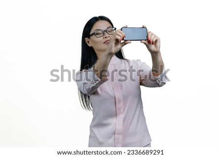 Young asian nerdy woman taking a horizontal selfie on her smartphone. Holding phone with both hands. Isolated on white background.