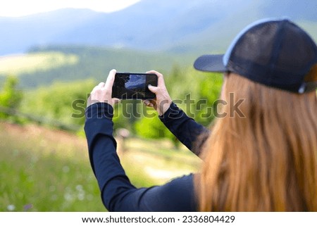 Woman taking a photo of a mountain landscape with their phone. The person is standing in a grassy field with wildflowers.