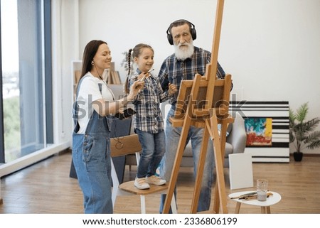Modern pensioner in headphones and smiling lady with wired earbuds dancing with cute girl while working on canvas. Artistic family finding inspiration in music while creating picture in living room.