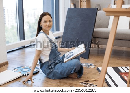 Portrait of adorable young female sitting back to large panoramic windows and posing on camera with picture. Creative artist demonstrating unfinished image of man on white canvas.