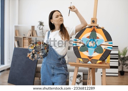 Pensive caucasian woman touching chin with paintbrush and leaning with elbow on wooden easel indoors. Pretty female holding colored palette and thinking about ideas for new artworks.