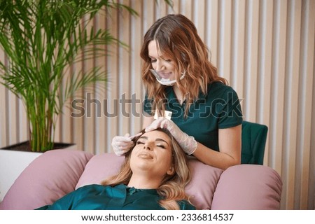 Beauty specialist comping client eyelashes after extension procedure. Female eyelashes technician in protective face mask combing woman lashes with disposable mascara brush in beauty salon. Royalty-Free Stock Photo #2336814537