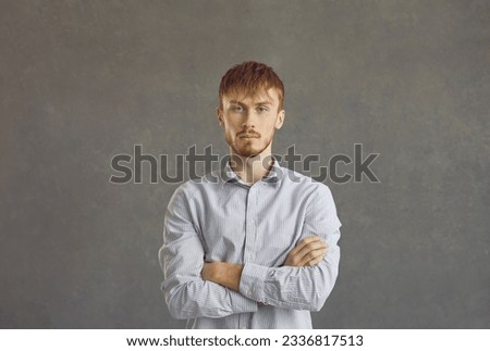 Serious young ginger guy in his 20s. Studio head shot portrait of man in his twenties with unsociable reserved introverted personality looking at camera standing arms folded against grey background Royalty-Free Stock Photo #2336817513