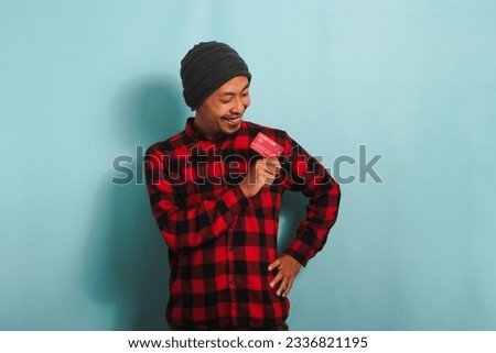 Excited young Asian man with a beanie hat and red plaid flannel shirt is showing a bank credit cards in his hands while standing against a blue background