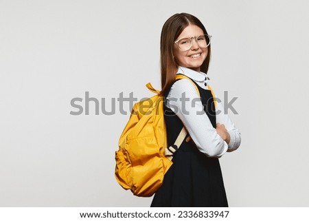 Side view of happy schoolgirl wearing glasses and yellow backpack holding crossed hands while smiling against white backdrop with free space for text Royalty-Free Stock Photo #2336833947