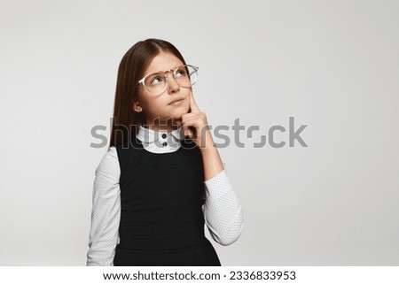 Nerdy schoolgirl wearing eyeglasses and uniform rubbing chin and looking away while thinking near empty space against white background