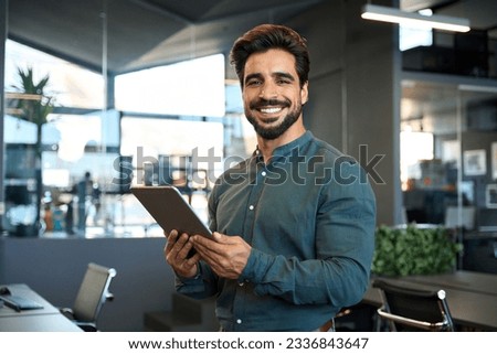 Smiling handsome young Latin business man entrepreneur using tablet standing in office at work. Happy male professional executive ceo manager holding tab computer looking at camera, portrait.