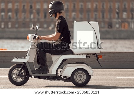 A man on a white electric scooter is in a hurry to complete the order. The courier carries the goods on a moped with a trunk along the river embankment. The transport company delivers the goods. Royalty-Free Stock Photo #2336853121