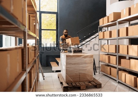 Employee wearing protective overall working in warehouse, carrying cardboard box and suitcase during small business inventory. African american supervisor preparing clients merchandise order Royalty-Free Stock Photo #2336865533