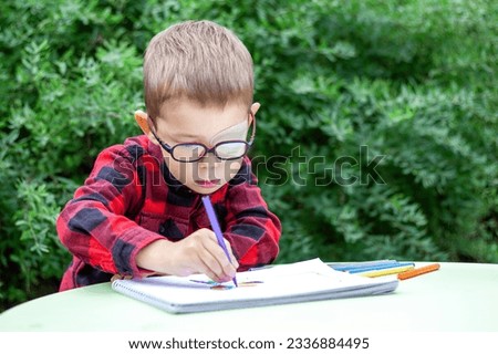 A little boy draws in the garden (outdoors). He wears glasses and an eye patch (occluder) to prevent amblyopia and strabismus (squint, lazy eye). The school students vision diseases problem. Royalty-Free Stock Photo #2336884495