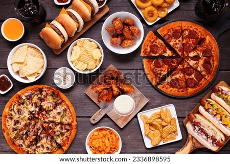 Junk food table scene. Pizza, hamburgers, hot dogs, chicken wings and salty snacks. Overhead view over a dark wood background.