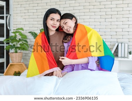 Asian young beautiful pretty female lesbian girlfriends couple love in casual outfits sitting posing on bed in bedroom smiling together covered with rainbow equality freedom LGBTQ gay proud pride flag