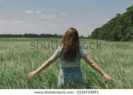 A woman enjoys the fresh air in nature in a green barley field. Summer countryside and gathering flowers. Atmospheric tranquil moment. Back view. Royalty-Free Stock Photo #2336934891