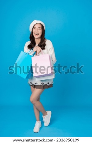 Full body shot of female online shopper female asian shopper with long hair, smiling, excited and holding many shopping bags, wearing hat, coat, skirt, photographed in studio, blue sky background.