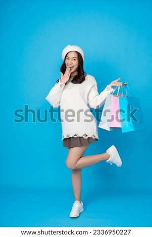 Full body shot of online shopper female Asian shopper with long hair excited and raised hands and legs posing. holding shopping bags wearing a hat coat skirt taking pictures in the studio blue scene