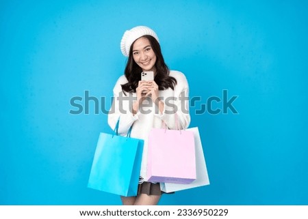 Online seller, long haired Asian female shopper Using a smartphone to place an order, smiling and having several shopping bags, wearing a hat, coat, skirt, taking pictures in the studio, blue scene
