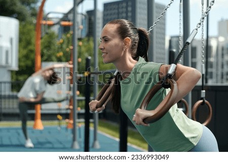 Happy young brunette sportswoman hanging on still rings on sports ground while working out against another woman doing exercise