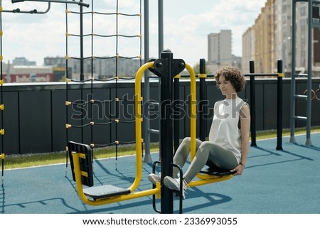 Young brunette sportswoman in activewear sitting on sports facilities while doing physical exercises in open air against urban environment Royalty-Free Stock Photo #2336993055