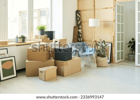 Stack of boxes, pictures, green domestic plants and armchair standing by large window of living room of new apartment during relocation
