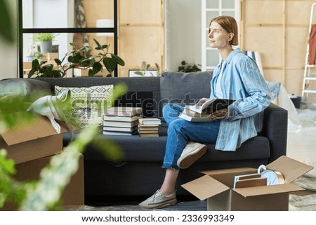 Young woman with stack of books on her knees sitting on soft comfortable couch in living room and having rest after relocation to new apartment Royalty-Free Stock Photo #2336993349