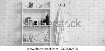 Shelving unit with supplies and bathrobe hanging on white brick wall in bathroom. Banner for design
