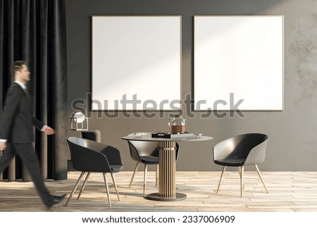 Businessman walking by two blank white posters on brown wall background with place for your logo brand or advertisement in sunlit living room with modern chairs around table on wooden floor, mockup