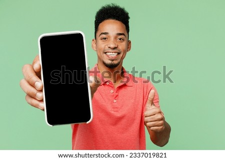 Young happy man of African American ethnicity he wears pink t-shirt hold in hand use mobile cell phone with blank screen workspace area show thumb up isolated on plain pastel light green background