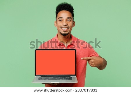 Young IT man of African American ethnicity wears pink t-shirt hold use work point finger on laptop pc computer with blank screen workspace area isolated on plain green background. Lifestyle concept