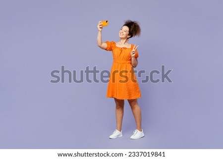 Full body young latin woman she wear orange blouse casual clothes doing selfie shot on mobile cell phone show v-sign isolated on plain pastel light purple background studio portrait. Lifestyle concept