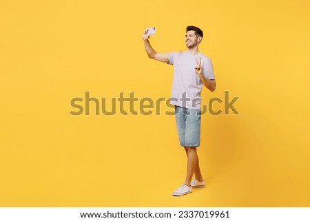 Full body fun young man wear light purple t-shirt casual clothes doing selfie shot on mobile cell phone post photo on social network show v-sign isolated on plain yellow background. Lifestyle concept