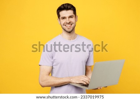 Young smiling cheerful happy fun caucasian IT man he wear light purple t-shirt casual clothes hold use work on laptop pc computer isolated on plain yellow background studio portrait. Lifestyle concept
